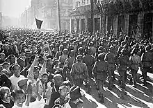 Photographie en noir et blanc d'une armée qui pénètre dans une ville, acclamée par la foule.