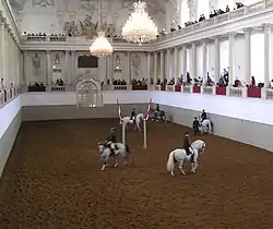 Dans une salle intérieure pourvue de galeries, richement décorée de marbre et de lustres, plusieurs chevaux et cavaliers en tenue effectuent une présentation.