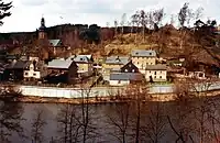 Photographie d'un petit village avec des maisons et une église situé dans une vallée boisée avec une rivière au premier-plan. Un imposant mur de béton sépare le village de la rivière.