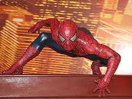 Statue de cire reproduisant le personnage Spiderman accroupi et légèrement assis sur sa jambe gauche, le buste en avant, avec la main gauche en avant et vers le bas. En arrière plan, on aperçoit des lumières de gratte-ciels, qui laissent supposer que Spiderman est sur un toit.