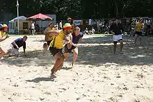 Sur du sable, un oueur en jaune, portant le ballon, est poursuivi par un autre joueur.