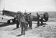 Groupe d'une dizaine d'hommes rassemblés autour d'un avion posé dans le désert.