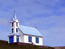 Église aux murs blancs et toit bleu, à flanc de butte, se découpant sur le ciel.