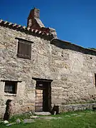 Vue d'un édifice médiéval en pierres, partagé en deux parties : à gauche, une maison, au toit de tuiles ; à droite, séparé par un petit clocher-mur, la chapelle, au toit couvert de lauzes.