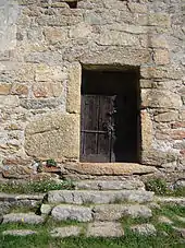 Porte de bois à deux battants, percée dans un mur de moellons très inégaux, à laquelle on accède par cinq marches de pierre.
