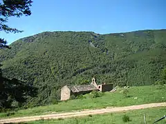 Photographie d'une chapelle de pierres au premier plan, située au flanc d'une vallée de montagne arborée.