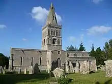 Vue de l'extérieur d'une église avec un clocher au centre de la nef, des pierres tombales se dressent au premier plan.