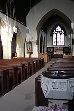 Intérieur d'une petite église simple, éclairé par un rayon de soleil. Un bénitier en pierre sculpté comportant une inscription illisible est aperçu au premier plan, sur la droite de l'allée centrale, devant les rangées de bancs en bois