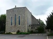 Photo d'un bâtiment à la façade de pierre grise imposante, percée de trois fenêtres en ogive, surplombé d'une croix chrétienne