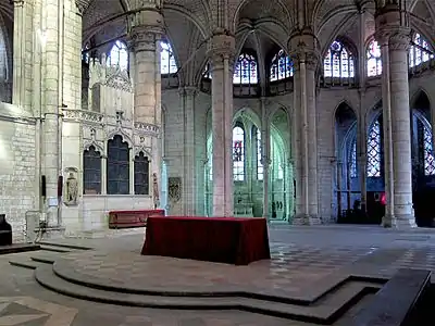 Autel avec une nappe rouge dans une grande pièce vide. Au fond de l'image, 4 ouvertures délimitées par 5 gros piliers donnent accès à des chapelles.