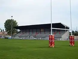 Vue d'un petit stade de rugby. Des poteaux de rugby et la pelouse au premier plan ; une tribune couverte en béton et un projecteur sur sa droite en arrière-plan