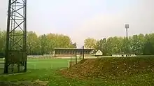 Tribune d'un terrain de football en herbe, derrière un pied de mat d'éclairage et une butte de terre.
