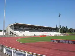 Vue d'une tribune couverte en béton, une piste d'athlétisme borde un terrain.