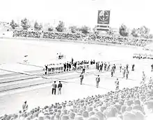 Photo en noir et blanc d'un avant-match de football, prise depuis les tribunes, avec le terrain au second plan
