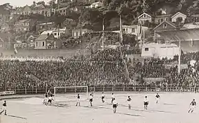 Photographie en noir et blanc d'un stade de football. Près du but, il y a plusieurs joueurs. Les tribunes sont remplies