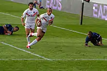 Joueur courant avec le ballon dans une main, suivi par un coéquipier, avec deux adversaires au sol.