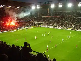 Photo montrant une vue d'ensemble d'un stade, avec des joueurs courant sur la pelouse, et un fumigène allumé dans une tribune.