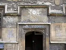 Une arche de pierre, sans porte ; au-dessus, les chiffres romains "VI" dans un cercle blanc et, à la craie, deux  avirons avec des pales vertes, les mots "Blades", "2001", "Men's 1st Torpid" ; à gauche, l'inscription "Balliol SEH Keble Merton", à droite, un dragon debout tenant un drapeau au-dessus des initiales «JCBC"