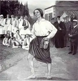 Une femme avec une chemise et une jupe regarde vers la gauche, tenant ses chaussures dans la main gauche.