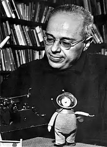 Portrait photographique en noir et blanc d'un homme souriant portant des lunettes avec en arrière-plan un mur tapissé de livres.