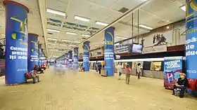Quai central avec des voyageurs et une rame à droite. le sol est jaune les colonnes recouvertes de publicités à dominante bleue et le plafond est blanc,