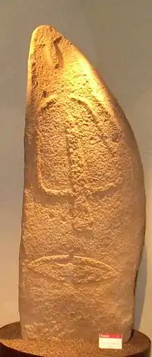 Menhir en pierre blanchâtre éclairé au-dessus par une lumière jaune, une palme et d'autres symboles en relief dessus.