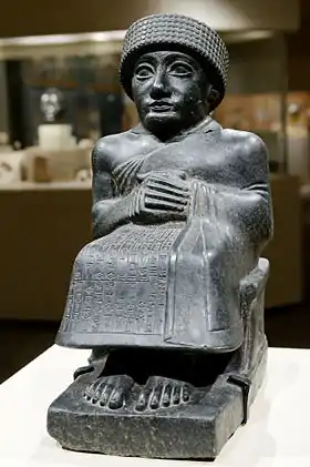 Statuette de pierre noire représentant un homme accroupi, vêtu d'une tunique longue et d'un bonnet.