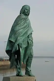 Vue d'une statue en bronze d’un homme en cape, sur fond marin.