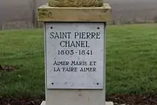 Plaque fixée sur le socle d'une statue (non visible), où est gravé en majuscules "Saint Pierre Chanel 1803-1841 Aimer Marie et la faire aimer".