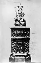 Photographie en noir et blanc d'une statue de la Vierge couronnée, avec un sceptre et portant Jésus enfant, sur un piédestal au treillis de bois.