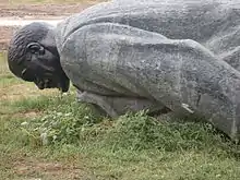 Photo couleur d'une statue de pierre grise (tête vue de profil et haut du corps d'un homme) renversée sur l'herbe verte d'un terrain vague.