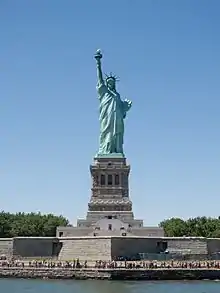 Photographie montrant la Statue de la Liberté derrière un drapeau américain, prise depuis un bateau touristique à New York Harbor.