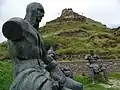 Trois statues assises représentant en gris des hommes en armure devant la forteresse de Gori en pierres de taille grises surplombant une colline d'aspect rocheux.