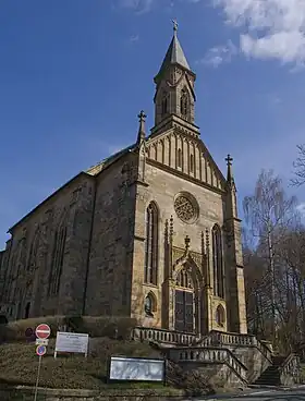 Photographie en couleurs de la façade d'une église néo-gothique de teinte beige et rosâtre, percée par deux oculi, une rosace et deux baies entourant l'entrée, sous un ciel azuré.