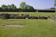 Photographie d'une pelouse où se dressent des bases de murs en ruine. Quatre petites stèles en briques, plus récentes, sont alignées au centre.