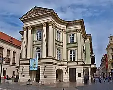 La façade d'un théâtre, à colonnade et fronton, entouré de rues piétonnes.