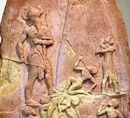 Photographie d'une partie d'une stèle sculptée en bas-relief. On y voit, à gauche, un grand personnage debout, coiffé d'un casque à deux cornes. Il marche vers la droite où se situent quatre petits personnages tombés ou suppliants. Au-dessus de ces-derniers est gravée une inscription en écriture cunéiforme.