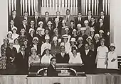 Photographie en noir et blanc d'un chœur placé devant un orgue.