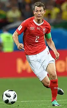 Photographie couleur d’un footballeur, de pied, en mouvement, le visage très expressif, vêtu d’un maillot rouge