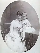 Tête contre tête Stéphanie assise sur un fauteuil porte une robe claire et esquisse un sourire en posant avec sa fille, souriante sous sa frange et assise sur une petite table.