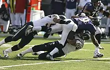 Un joueur de football américain en combinaison violette et blanche est tiré aux sols par deux joueurs en tenue blanche.