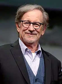 Un homme souriant d'une soixantaine d'années à la bouche fermée, aux cheveux courts et grisonnants, portant une barbe naissante grisonnante et une monture de lunettes noire.