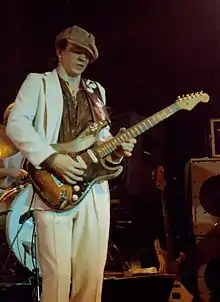 Photo d'un homme jouant de la guitare, vêtu d'une veste et d'un pantalon blancs