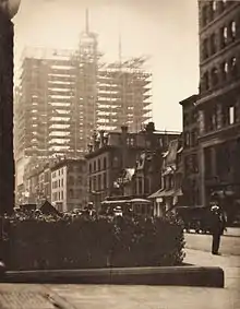 Photographie réalisée depuis une rue bordée de bâtiments traditionnels de quelques étages avec le squelette métallique d'un gratte-ciel à l'arrière-plan