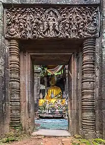 Porte en pierre avec des colonnes et des reliefs bouddhistes, menant à une pièce avec une statue de Bouddha assis vêtu d'une tunique orange, une table avec des pierres et des reliques bouddhistes, dans le complexe du temple hindou khmer de Wat Phou. Septembre 2019.