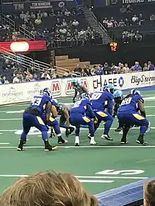 Les joueurs du Storm de Tampa Bay en uniforme bleu foncé, prêts pour le snap. Tribunes partiellement occupées à l'arrière plan.