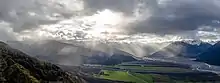 Une grande vallée où des rayons du Soleil sortis des nuages illuminent des champs et le cours d'eau ; tout est couronné par une chaîne de montagnes.