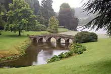Grand parc avec bois et pelouses. Un pont à arches de pierre franchit un bras de rivière