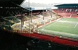 Une tribune de football avec des sièges verts, des piliers en son centre soutenant un toit qui s'incline au-dessus de la tribune.