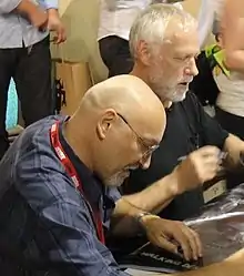 Deux hommes assis côte à côte pour une séance d'autographes.